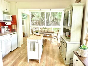 A kitchen with a dining area, wooden flooring, white cabinets, a microwave, and a large window with a forest view.