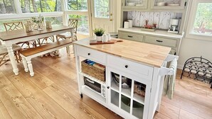 Une cuisine avec une table en bois, des meubles blancs et une vue sur les arbres à travers de grandes fenêtres.