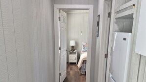 A narrow hallway with wooden floors, white walls, and a patterned rug. A white refrigerator is on the right, and a bed with a nightstand and lamp is visible in the distance. - Little Barnstead Tiny Home (Big Cabin)