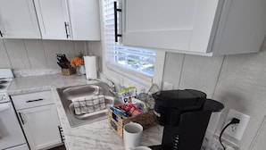 A modern kitchen with white cabinets, a marble countertop, a coffee maker, and a window with blinds. - Little Barnstead Tiny Home (Big Cabin)