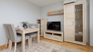 Una sala de estar moderna con una mesa de comedor de madera, sillas tapizadas grises, un televisor de pantalla plana, un mueble con puertas de cristal y una pequeña estantería con libros.