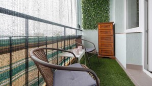 Un balcón con barandilla de malla, una pequeña mesa con sillas, un armario de madera y una pared verde.