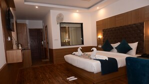 Habitación de hotel con piso de madera, una cama grande con ropa blanca, una silla azul, un armario de madera y una ventana con cortinas.