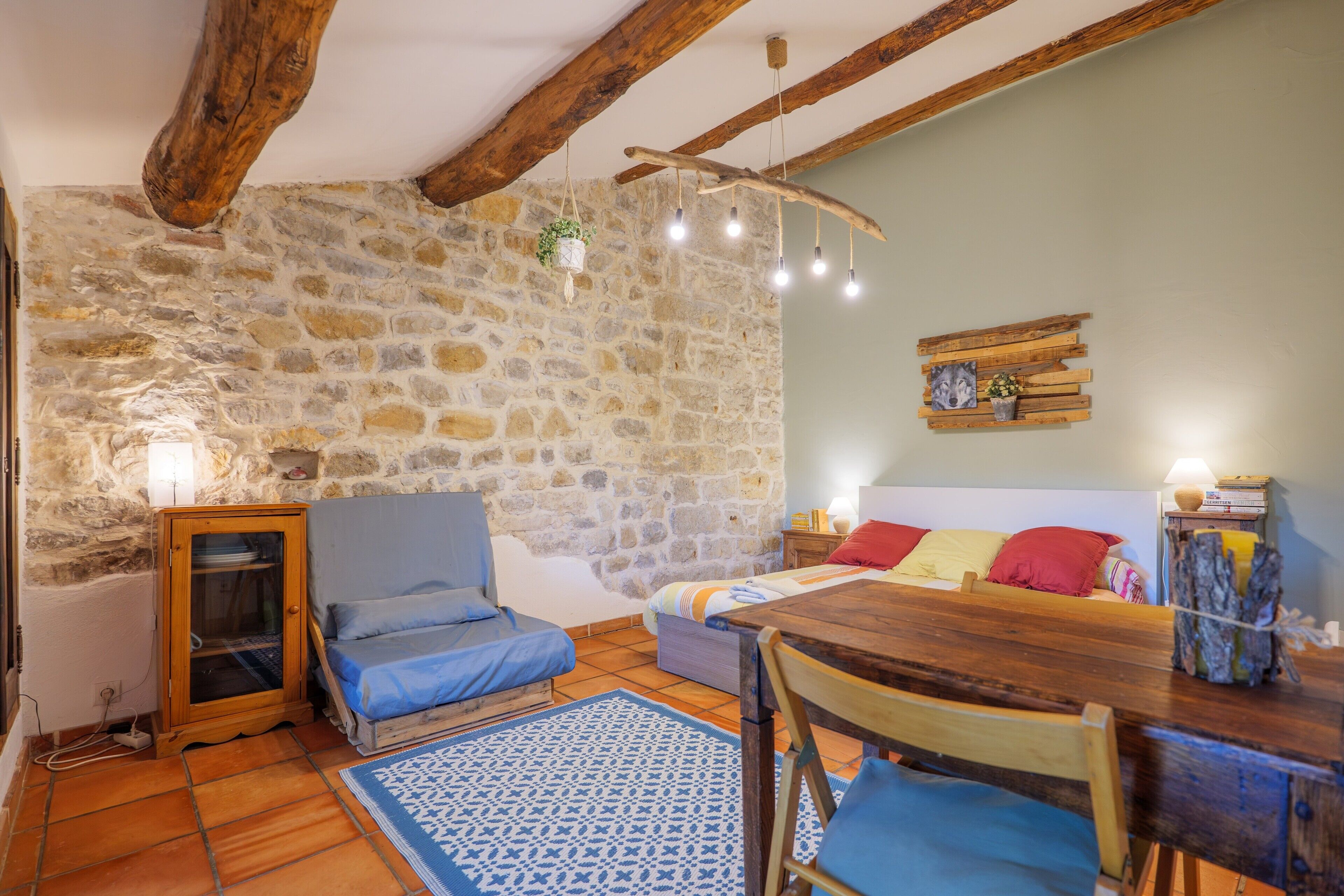 A rustic room with a stone wall, wooden beams, a bed with blue and red pillows, a wooden table, and a hanging light fixture.