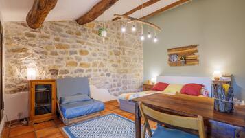 Une pièce rustique avec un mur en pierre, des poutres en bois, un lit agrémenté de coussins bleus et rouges, une table en bois et un luminaire suspendu.
