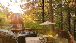 A wooden deck with a sofa, chairs, and a table, surrounded by trees and a cabin in the background. - Treehouse Retreat | Sleeps 10 | Big Deck (Shaver Lake)