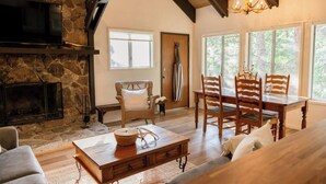 A cozy living room with a stone fireplace, wooden dining table, and comfortable seating. - Treehouse Retreat | Sleeps 10 | Big Deck (Shaver Lake)