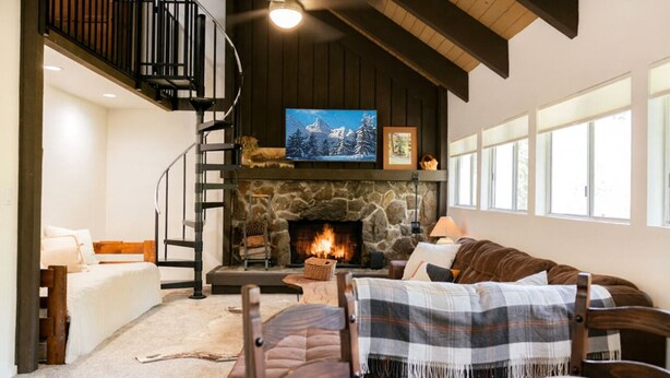 A cozy living room with a stone fireplace, a large TV mounted above it, and a comfortable seating area with a sofa and chairs. - Treehouse Retreat | Sleeps 10 | Big Deck (Shaver Lake)