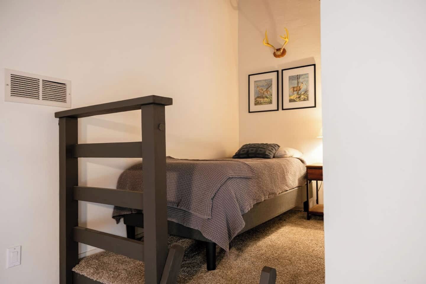 Un dormitorio con cama, cabecera, mesita de noche, lámpara y dos cuadros enmarcados en la pared.