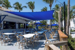 An outdoor seating area with tables and chairs, a bar, and palm trees in the background.