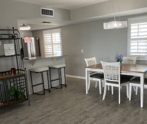 A dining area with a wooden table and white chairs, a bar area with stools, and a kitchen counter with appliances.