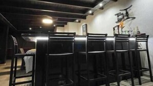 Una zona de bar con taburetes altos y una barra, un techo con vigas a la vista y una lámpara fijada a la pared.