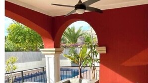 Un espacio de estar techado al aire libre con mobiliario de mimbre, ventilador de techo y vista a una piscina.