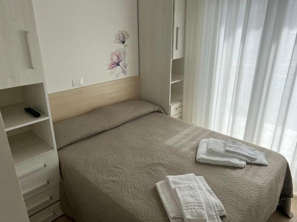 A neatly made bed with a beige quilt, a bedside table with a remote control, and a wall with a floral painting.