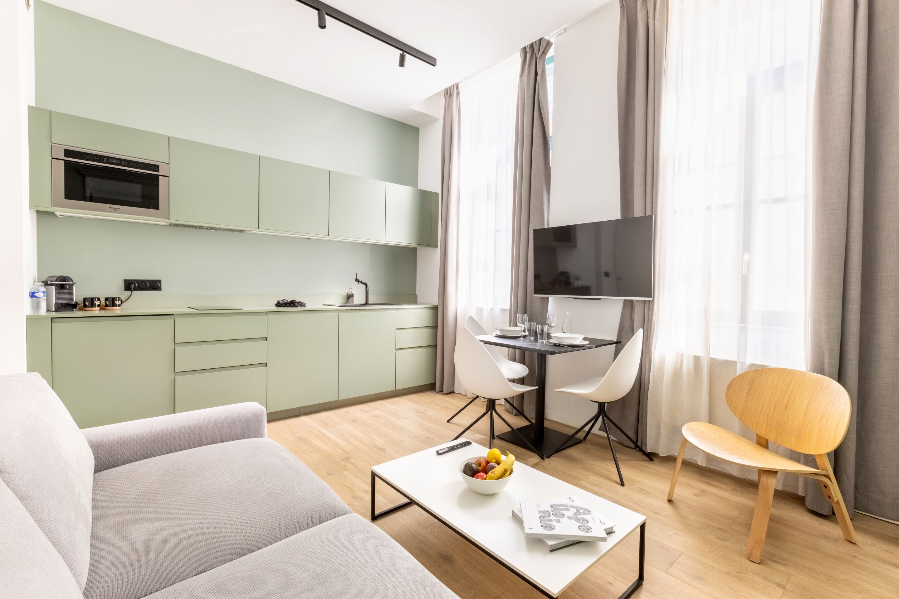 A modern living room with a sofa, a coffee table with a bowl of fruit, a dining area with a table and chairs, a kitchen with green cabinets and a microwave, and a TV mounted on the wall.
