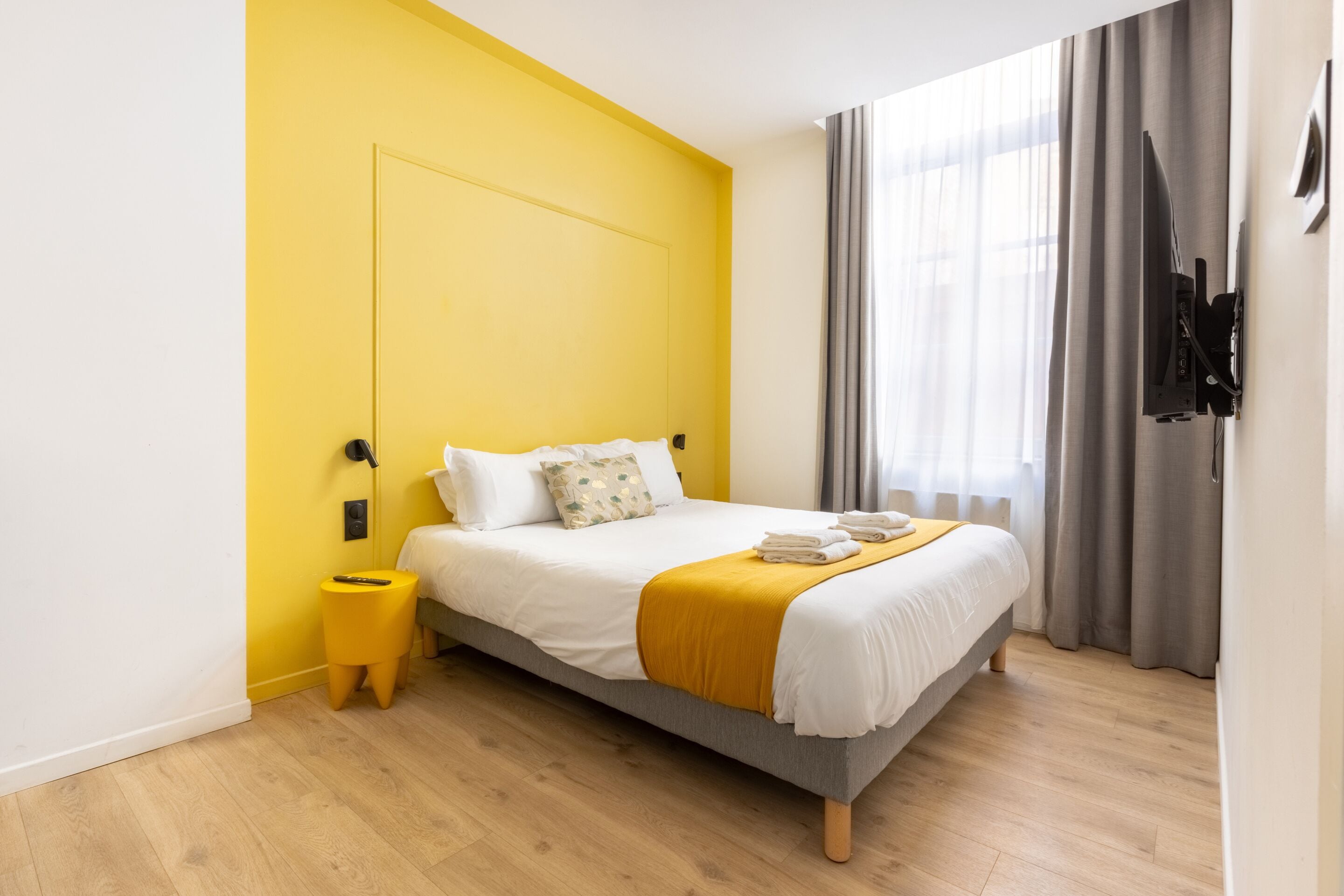Quarto de hotel moderno com cama, mesa de cabeceira, televisão na parede e janela com cortinas.