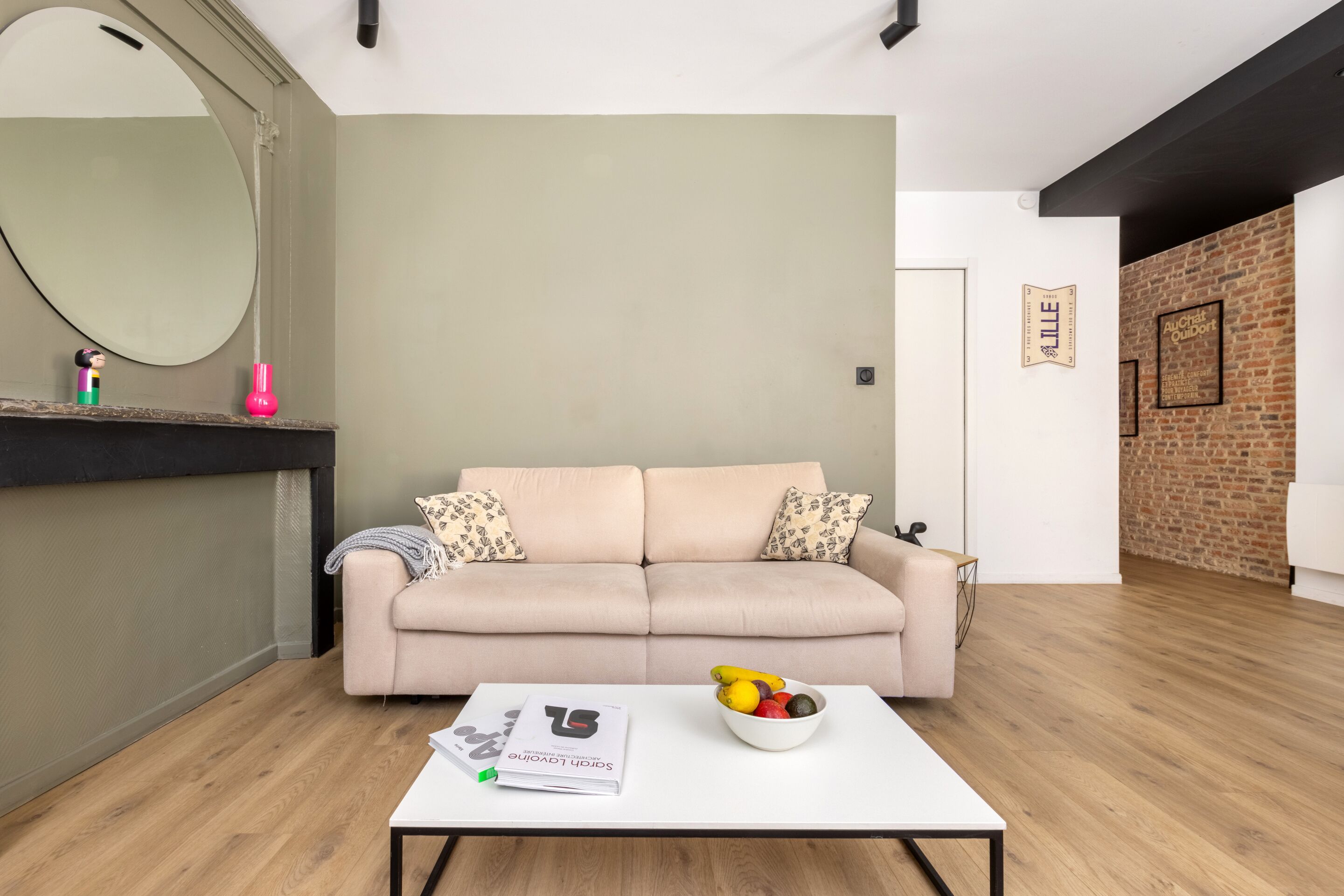 A modern living room with a beige sofa, a white coffee table with a bowl of fruit, a magazine, and a mirror on the wall.