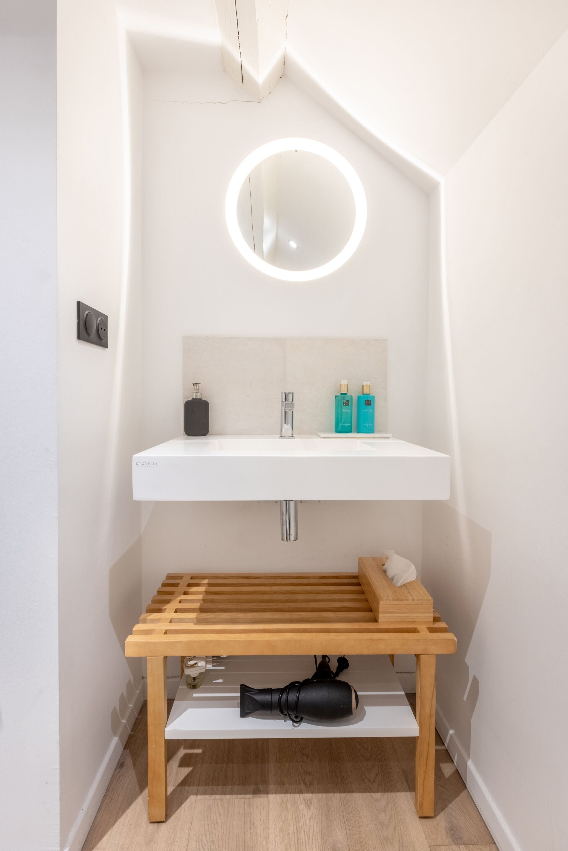 Un bany modern amb un rentapans blanc, un mirall amb llum circular i una cadira de fusta a la part inferior.