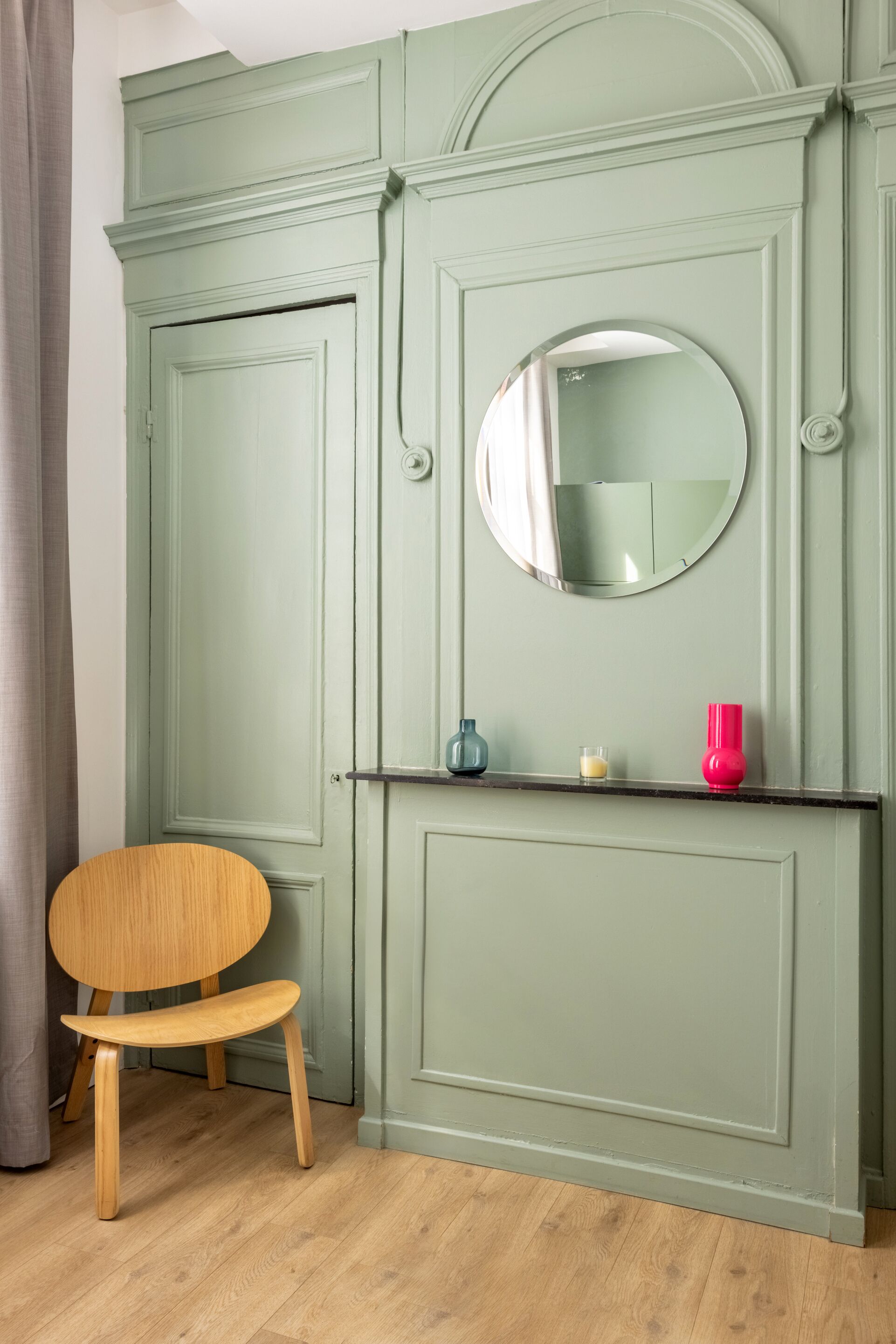 Um quarto com um guarda-roupa verde, um espelho redondo, uma cadeira de madeira e uma estante com um pequeno vaso e uma vela.