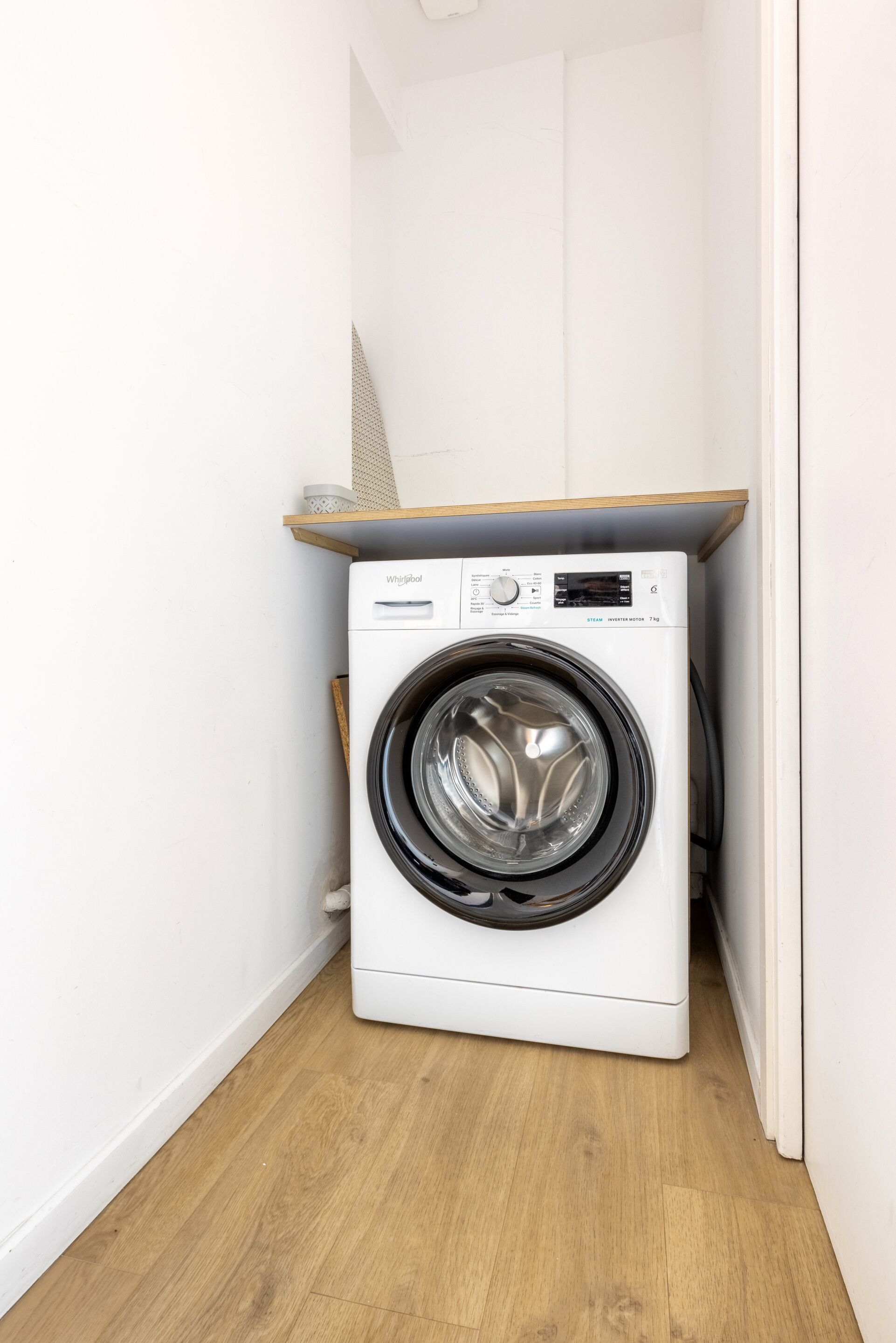 A compact, white front-loading washing machine in a small laundry room with wooden flooring and a white wall.