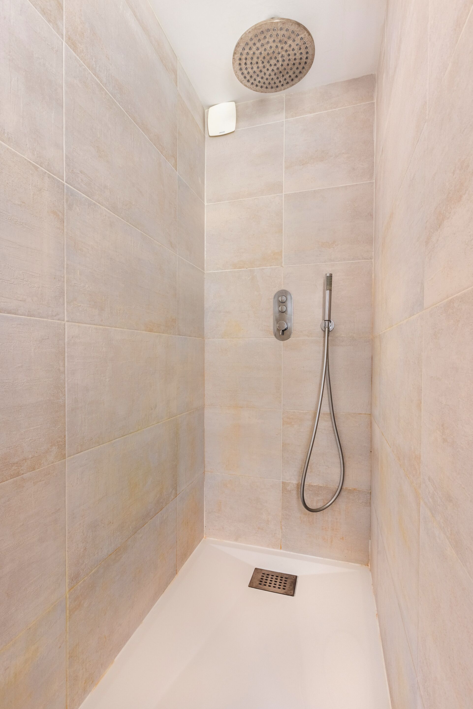 A shower with a handheld showerhead, a wall-mounted control panel, and a drain.