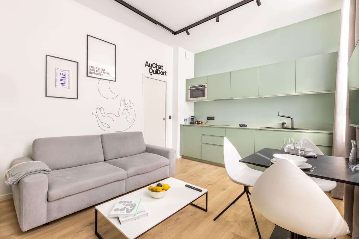 Un salon moderne avec un canapé gris, une table basse sur laquelle se trouve un saladier rempli de fruits, et un fauteuil blanc. On aperçoit, au fond, une cuisine avec des meubles verts et un four à micro-ondes.