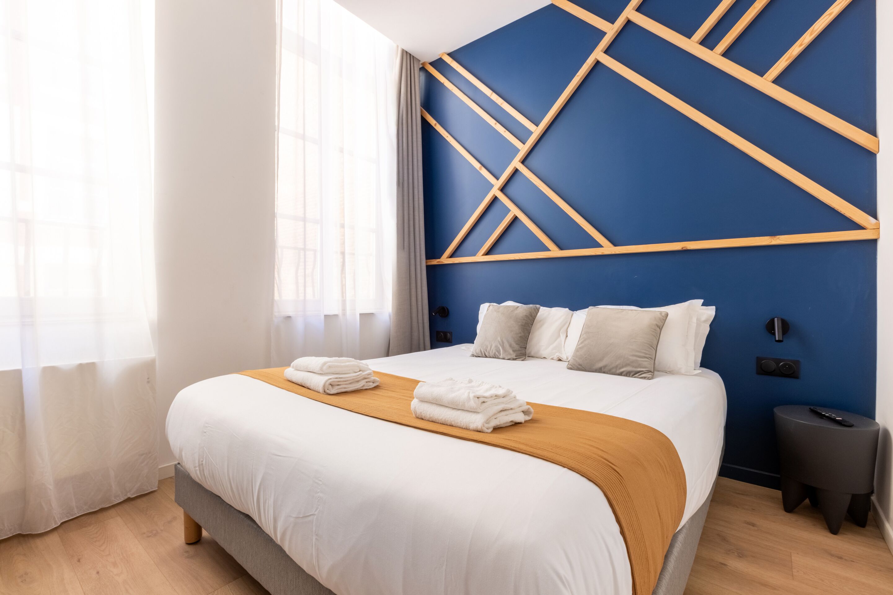 Una cama bien tendida con sábanas blancas y un cabecero de madera, una mesita de noche con un control remoto, y una ventana con cortinas de encaje.