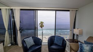 Una habitación con un gran ventanal corredero que da a vista de playa, dos sillones de cuero con cojines azules, un sofá marrón con cojines azules y una mesita de noche con una lámpara.