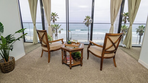 Una habitación con un ventanal grande con vista a la playa, dos sillas de madera, una mesita con una bandeja y una planta en maceta.