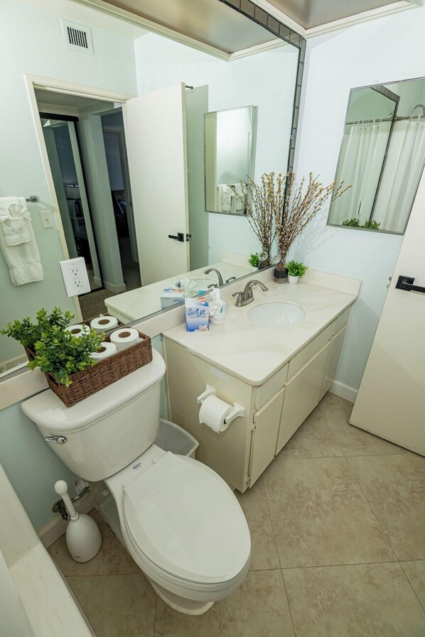 Un baño moderno con inodoro, lavamanos y espejo. Hay una cesta con papel higiénico y una planta sobre el inodoro.