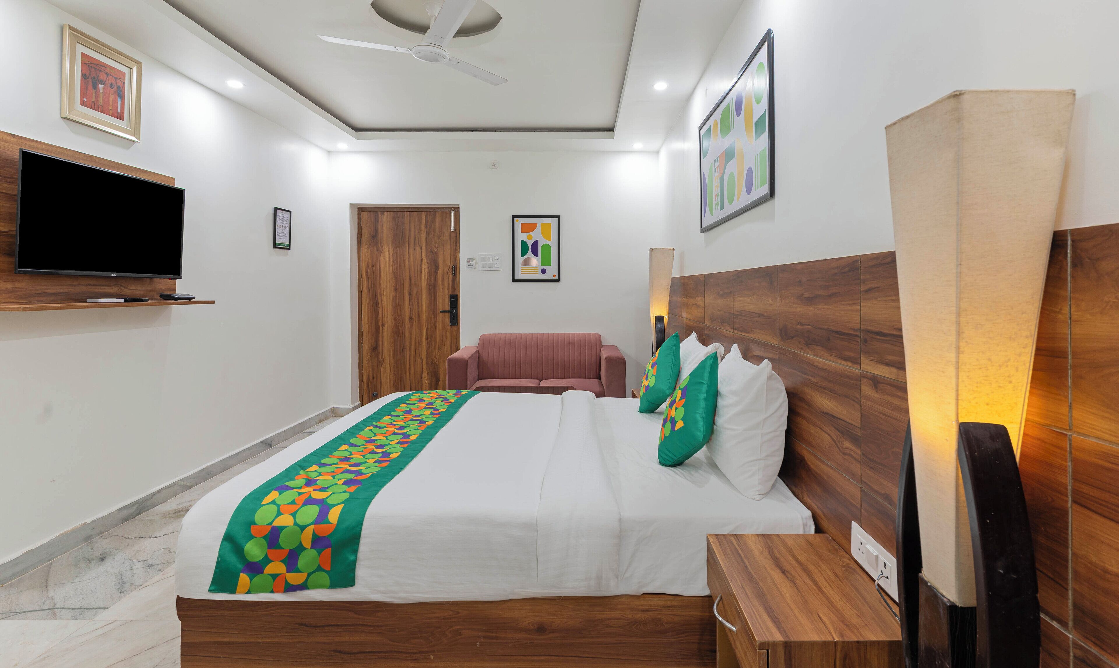 Una habitación de hotel con una cama, televisor montado en la pared, cabecera de madera y ventilador de techo.
