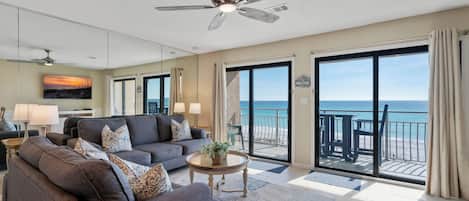 A spacious living room with a gray sofa, a coffee table, and a ceiling fan. A large window offers a view of the beach.
