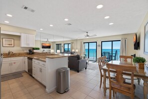 A modern kitchen with white cabinets, a granite countertop, and a dishwasher. There is a dining area with a wooden table and chairs, and a large window offering a view of the ocean. - Crystal Villas B9 New Listing Specials! Gulf Front, Pool, Free Beach Service (Destin)