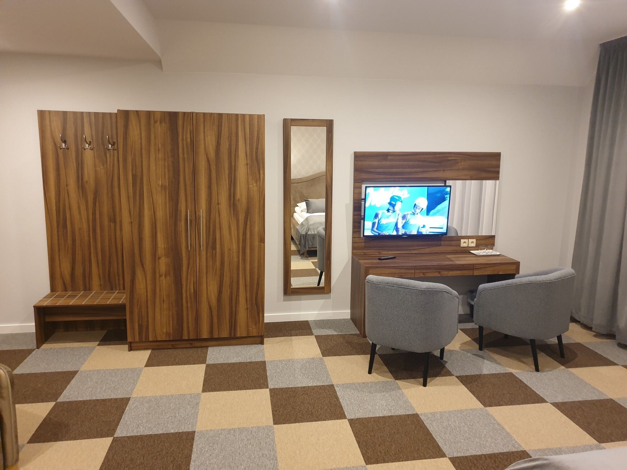 Quarto de hotel com guarda-roupa de madeira, uma escrivaninha com televisão, duas cadeiras cinzentas e chão xadrez.