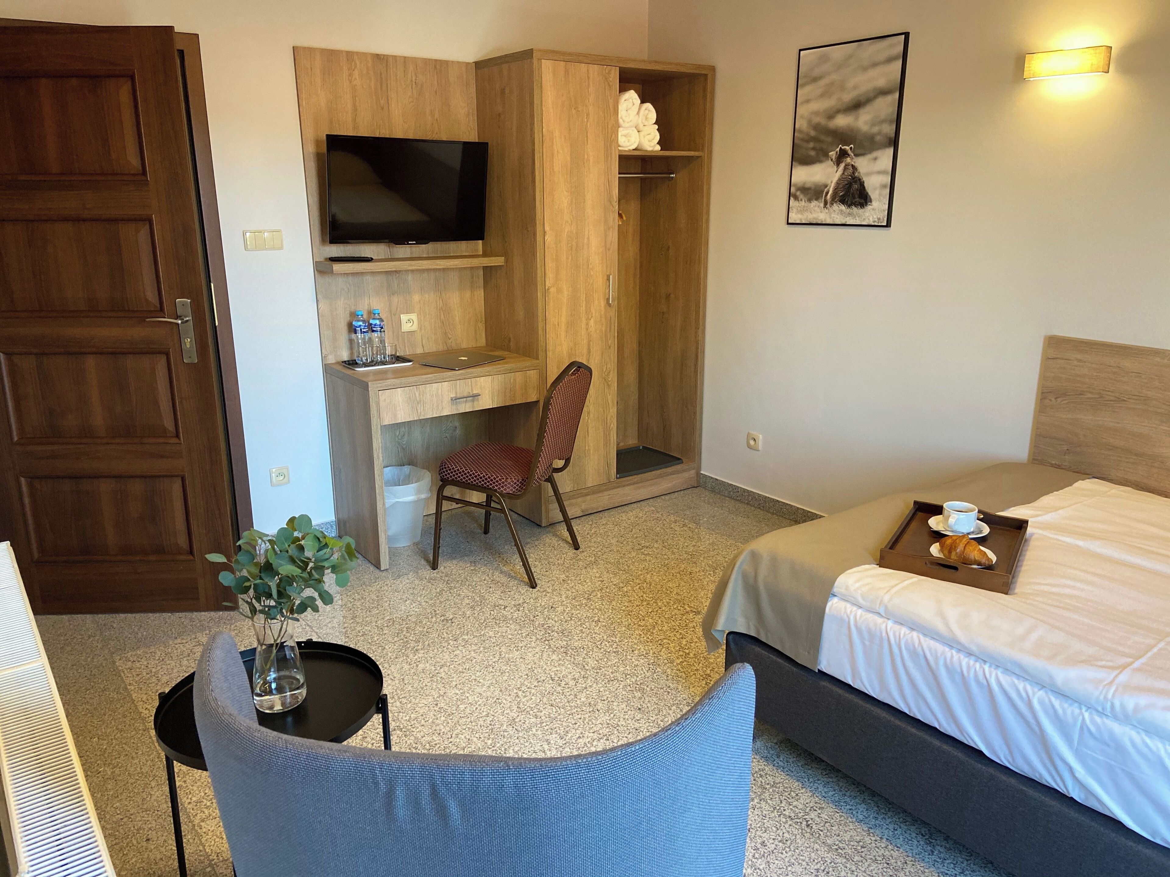 Habitación de hotel con cama, escritorio con computadora, silla, televisor, estante con toallas, cuadro enmarcado y bandeja con un croissant y una taza.