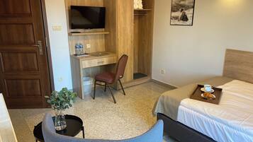 Habitación de hotel con cama, escritorio con computadora, silla, televisor, estante con toallas, cuadro enmarcado y bandeja con un croissant y una taza.
