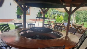 A round wooden table with a built-in grill, surrounded by wooden chairs, under a wooden pergola with hanging chains. - Azretreat (gmina Ostrowite)