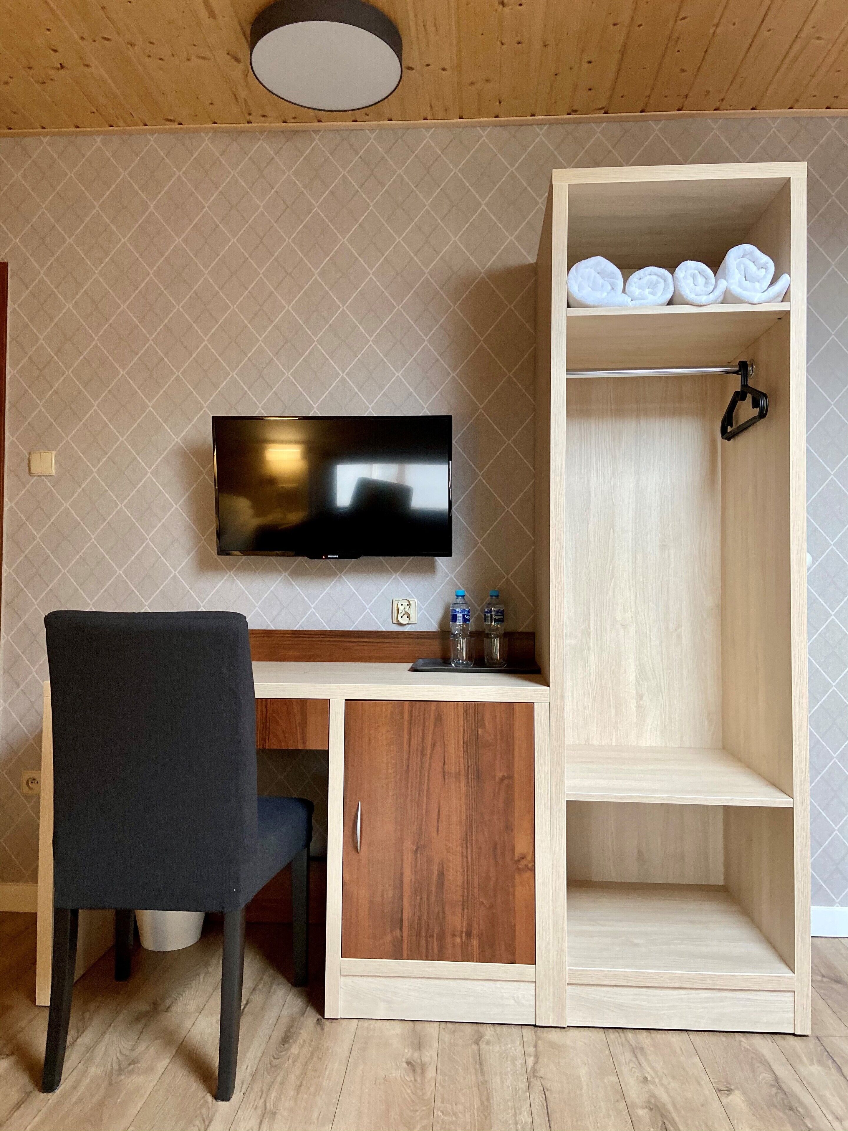 Una habitación con un escritorio de madera, una silla negra, un televisor de pantalla plana y un armario de madera con estantes.
