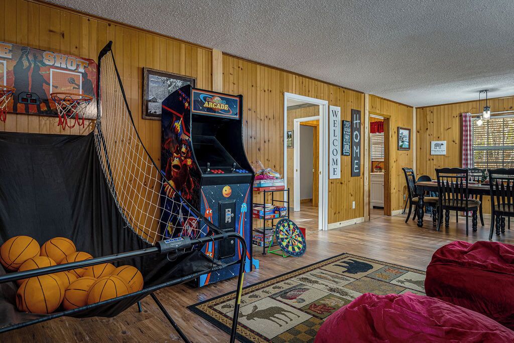 A room with a basketball hoop, an arcade game, a dartboard, and a dining area with a table and chairs.