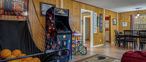 A room with a basketball hoop, an arcade game, a dartboard, and a dining area with a table and chairs.