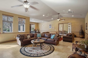 A spacious living room with a large brown leather sectional sofa, a coffee table, and a patterned rug. There are ceiling fans, recessed lighting, and large windows with blinds. - The Grand Retreat - Brand New! (Hurricane)