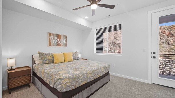 A bedroom with a bed, a nightstand, a ceiling fan, a window with blinds, and a door with a view of a rocky landscape. - The Grand Retreat - Brand New! (Hurricane)