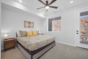 A bedroom with a bed, a nightstand, a ceiling fan, a window with blinds, and a door with a view of a rocky landscape.