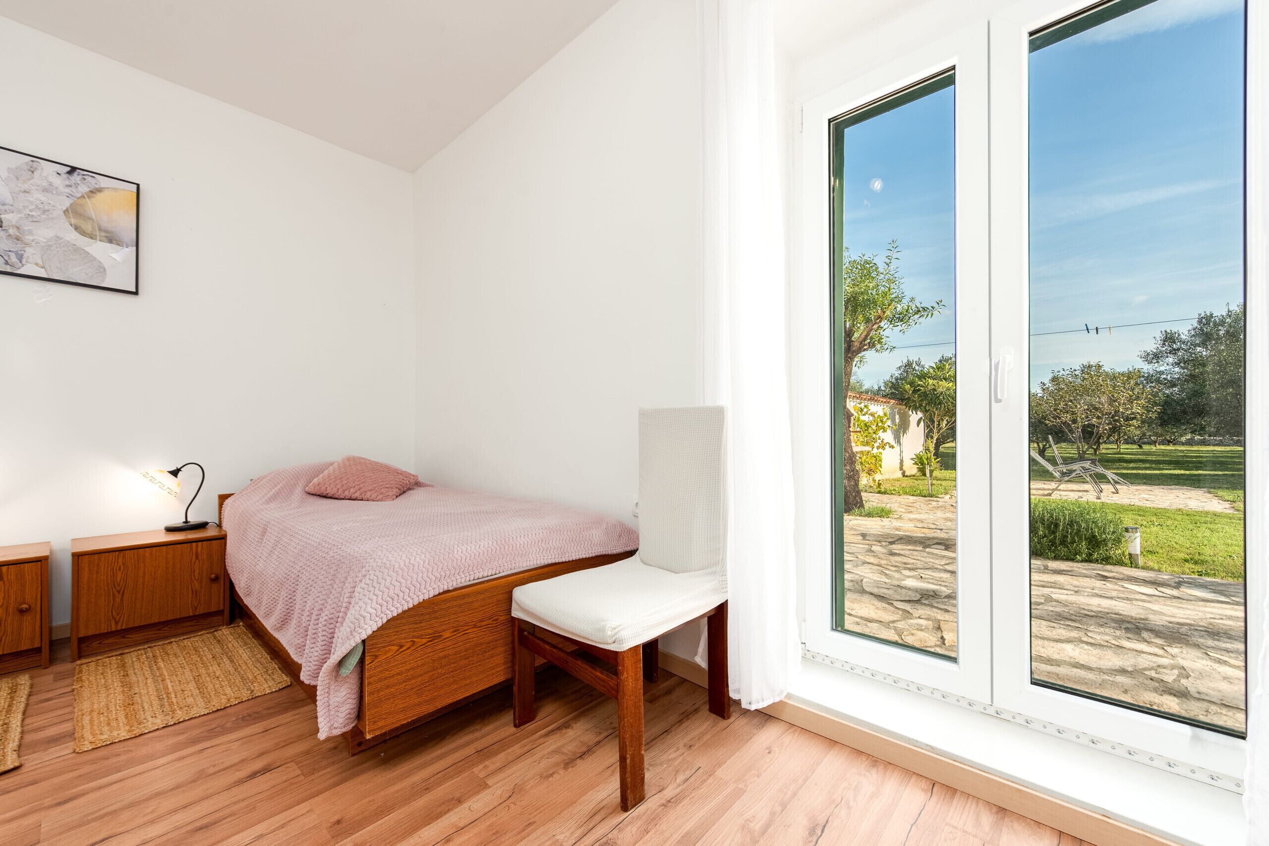 Un dormitorio con cama de madera, una mesita de noche, una silla, una ventana con vistas a un jardín y una lámpara de pared.