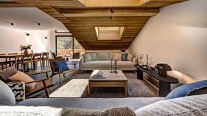 Ein gemütliches Loftsessel-Wohnzimmer mit hohen Decken, Holzbalken, einem Oberlicht, einer Couch, Sesseln, einem Couchtisch und einem kleinen Beistelltisch mit Kerzen.