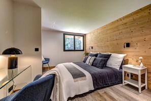 Een moderne slaapkamer met een houten accentmuur, een groot bed met veel kussens, een nachtkastje met een vaas en een raam met uitzicht op bomen.