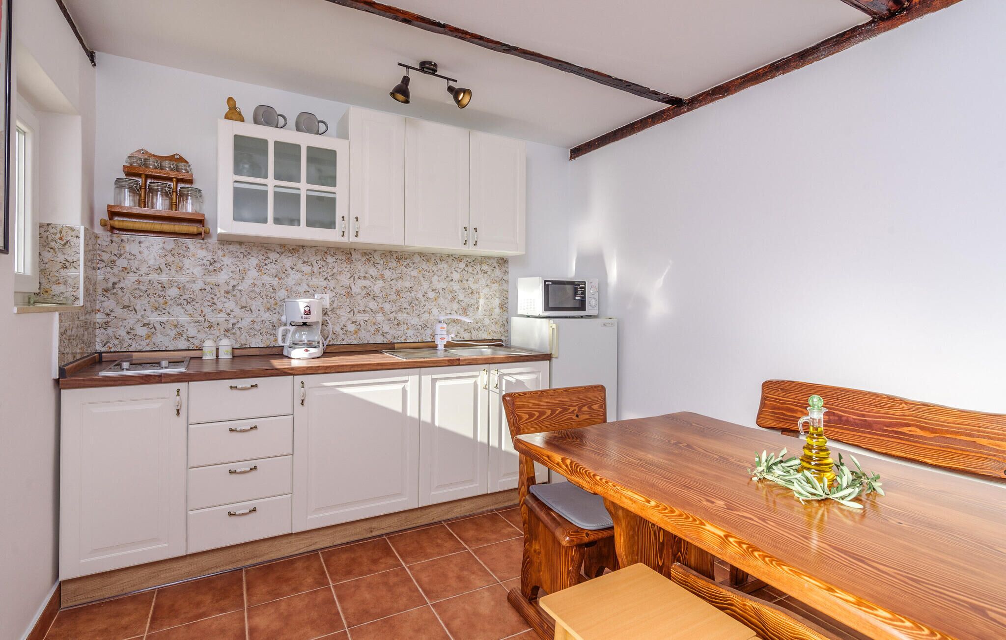 A compact kitchen with white cabinets, a coffee maker, a microwave, and a dining table with a wooden bench.