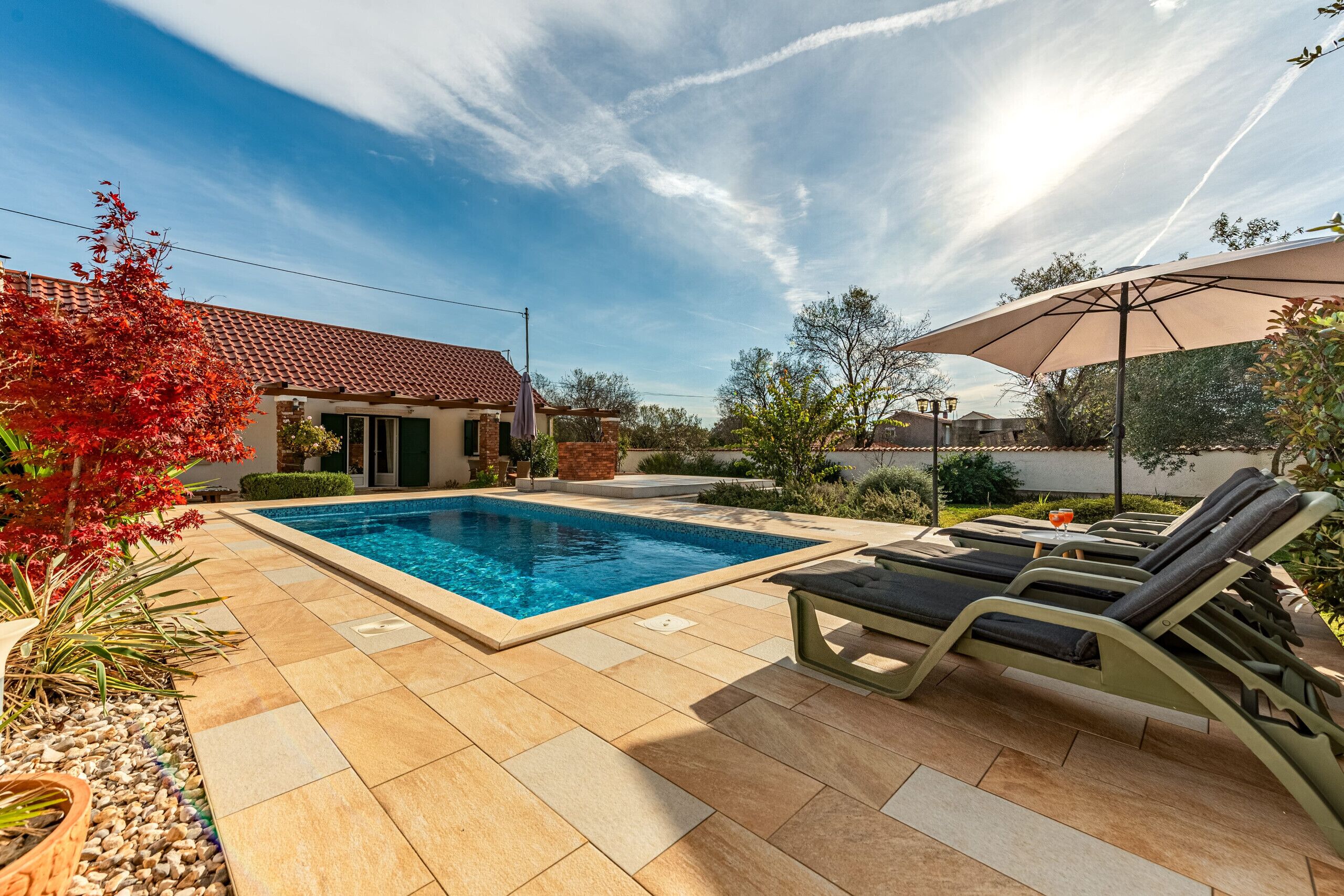 Ein Schwimmbecken mit Liegestühlen und einem Sonnenschirm, umgeben von einer Terrasse mit Fliesenboden und Pflanzen.