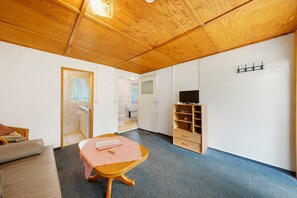 Ein Zimmer mit Holzbalkendecke, einem kleinen Tisch mit rosa Tischdecke, einer Couch, einem Fernseher auf einem Regal und einer Tür zu einem Badezimmer.