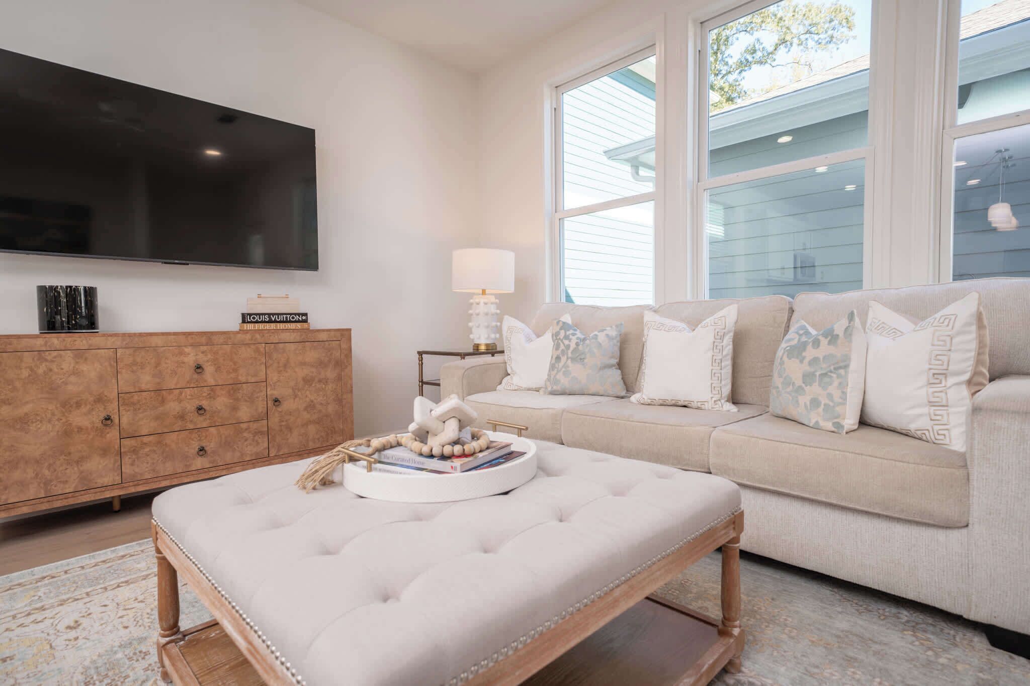 A living room with a large flat-screen TV mounted on the wall, a beige sofa with patterned cushions, a wooden coffee table with a decorative centerpiece, and a side table with a lamp.